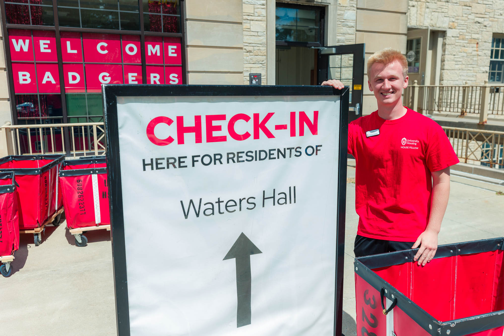 A person stands next to a sign reading "Check-In here for residents of Waters Hall" with a sign reading "Welcome Badgers" in the background
