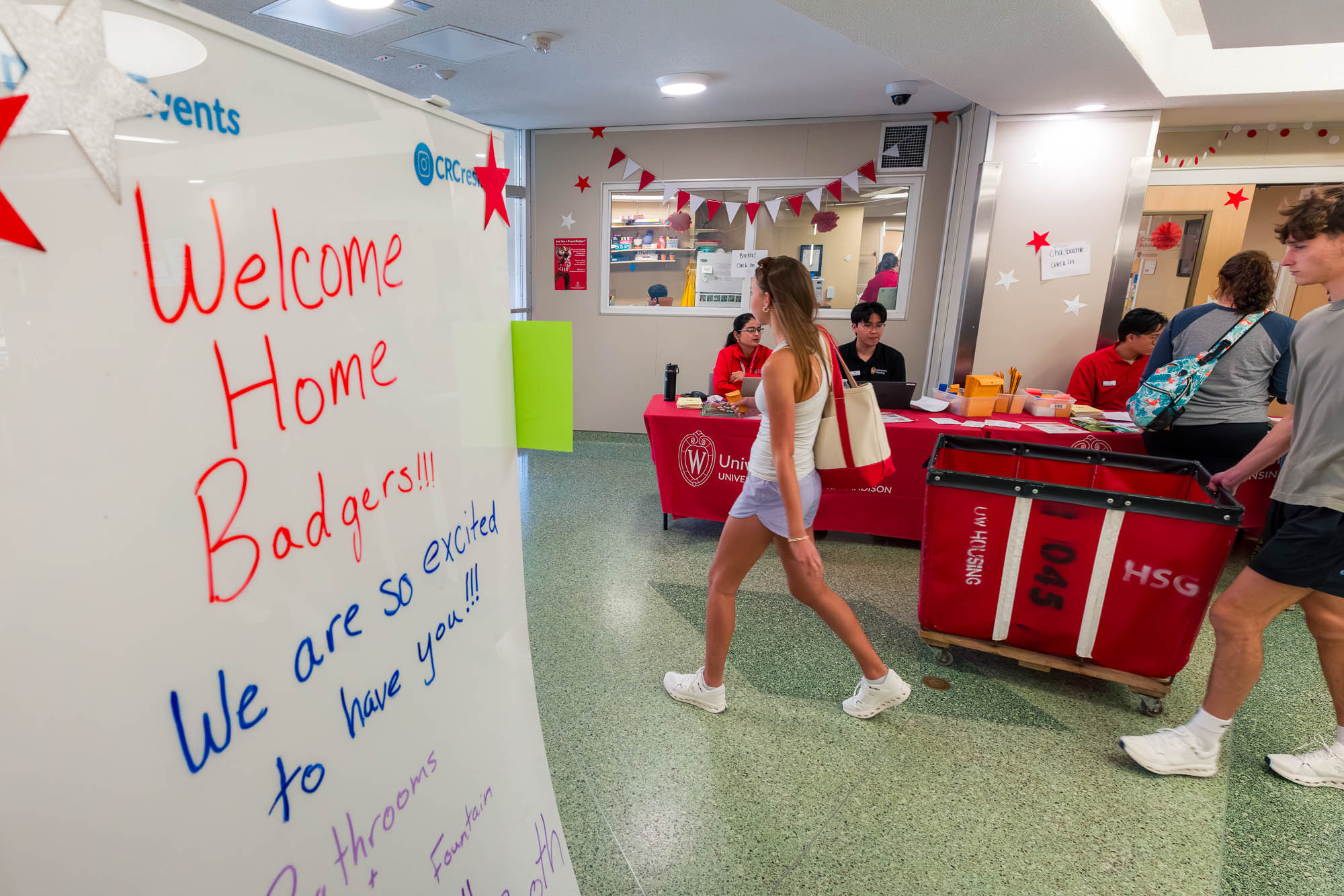 A sign reads "Welcome Home Badgers!!! We are so excited to have you!!!" while people walk in the background with a storage cart