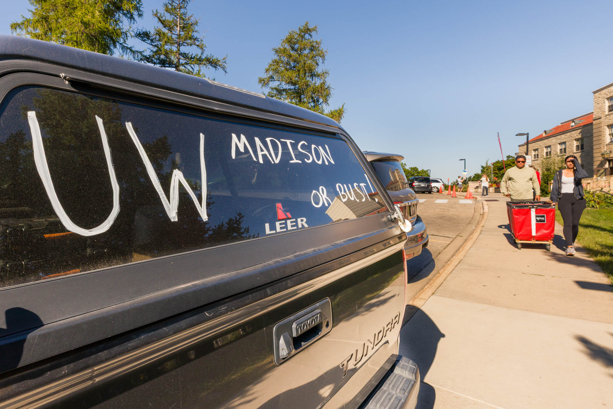 The rear window of a truck in the foreground with the words "UW–Madison or Bust!" written on it in marker with two people and a moving cart in the background