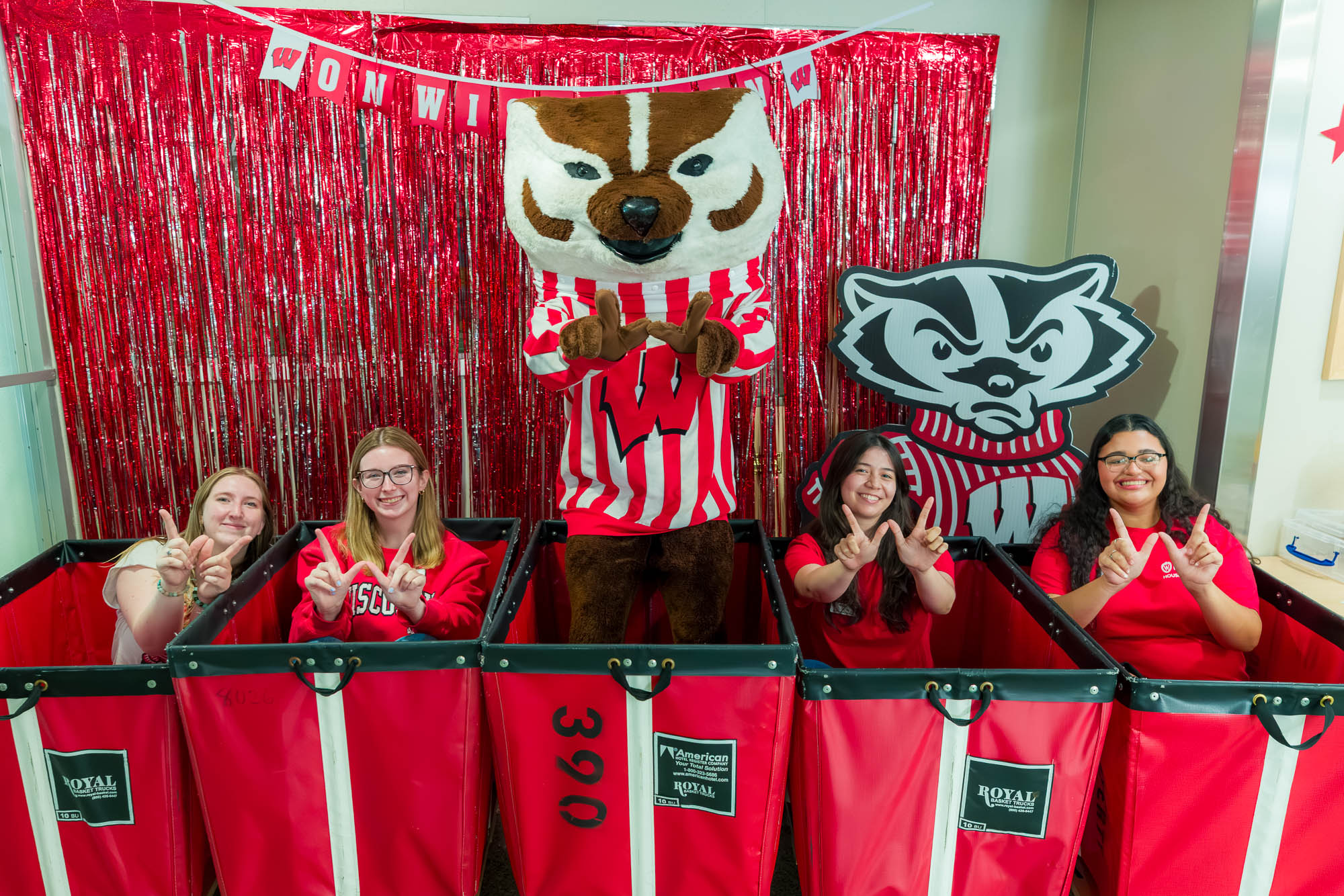 Four people sitting in moving carts making "W's" with their hands with Bucky Badger standing in a moving cart in the middle