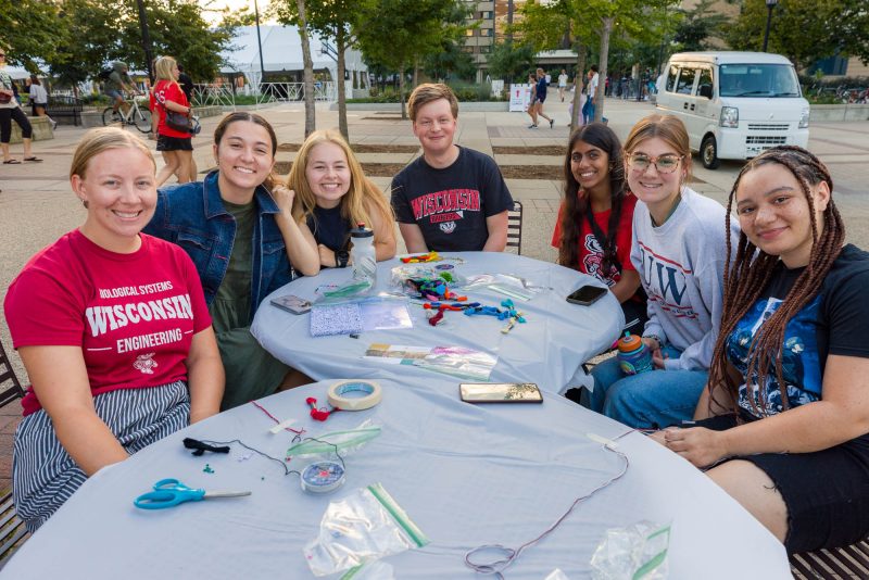 Seven people pose for a photo at tables outside while making bracelets