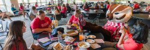 Three people sit at a table with Bucky Badger while he puts his feet up on the table