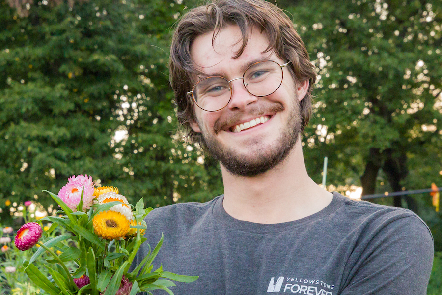 A student with glasses and facial hair smiles for a portrait while holding a colorful bouquet of flowers