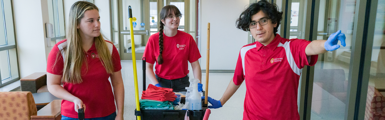 Three Facilities students in red shirts transporting custodial supplies down a hallway