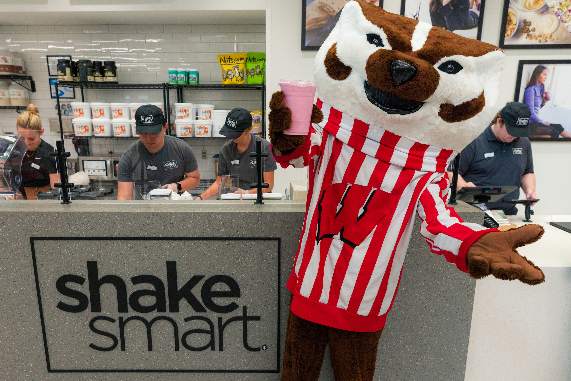 Bucky Badger holding up a pink protein shake while Shake Smart staff work behind the counter