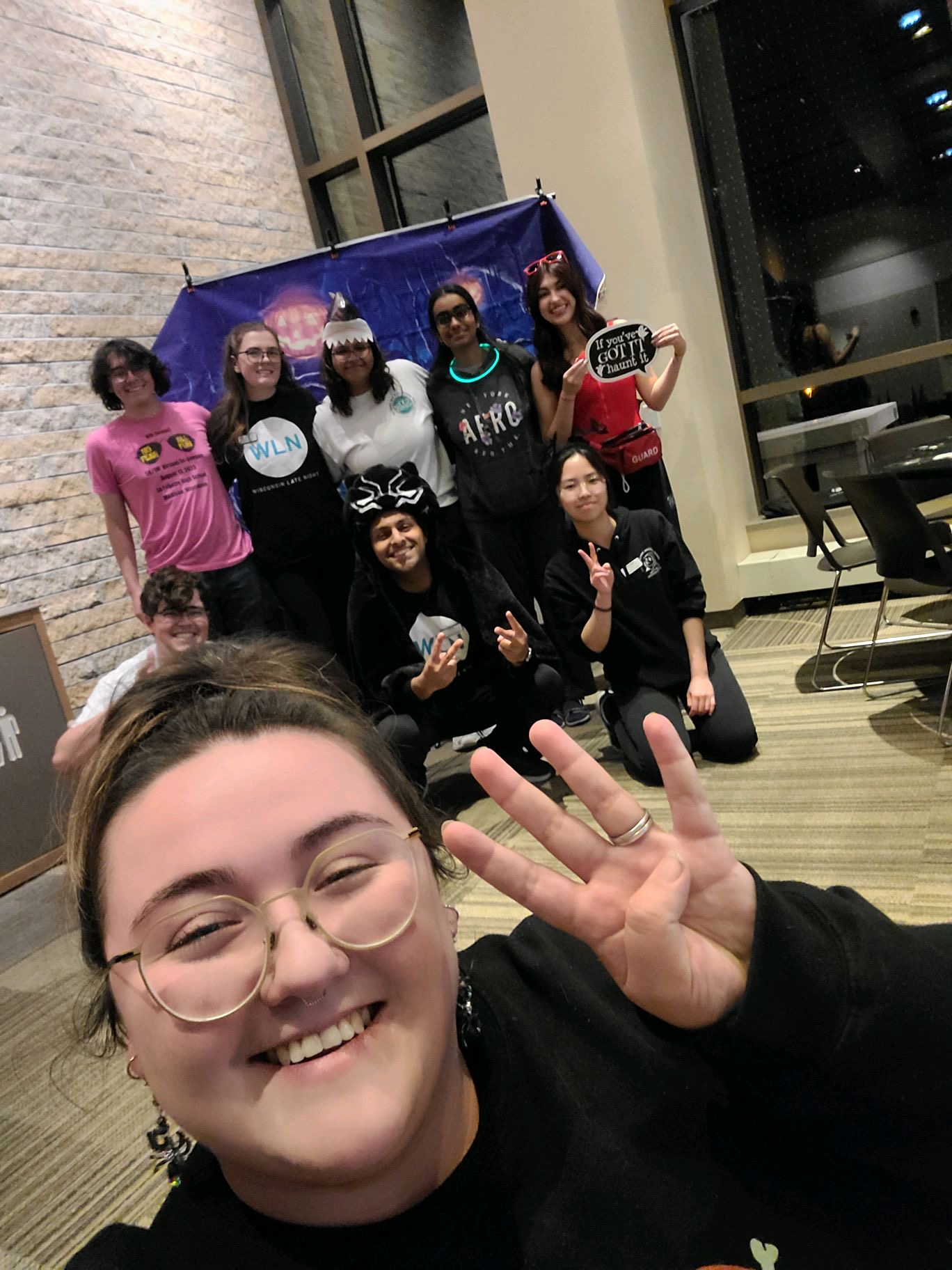 One women holding up four fingers and smiling for a selfie with eight others behind her