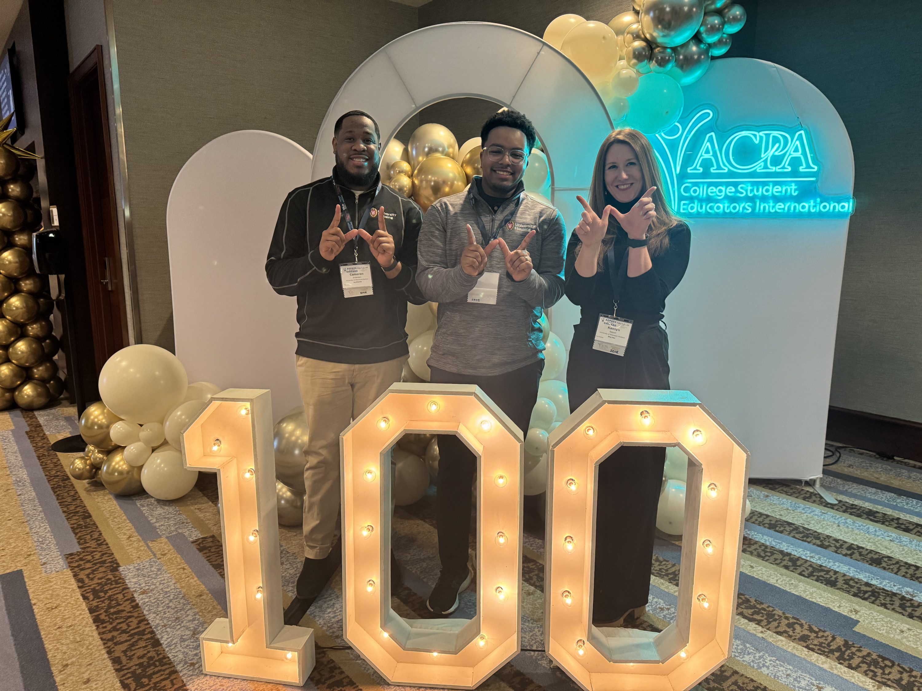 Three people smiling and making a motion "W" with hands standing behind a 100 lighted numbers