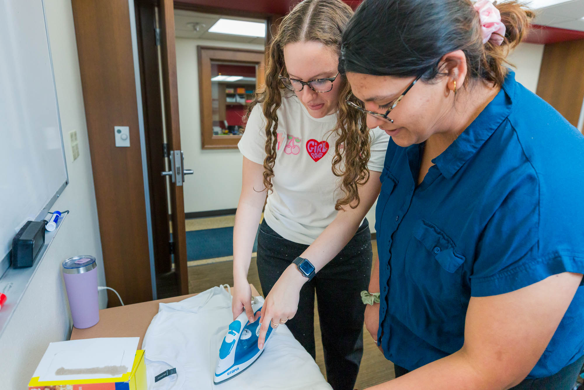Madison Eiler uses an iron to prepare shirts with another student employee in Carson Gulley Center for a Wisconsin Weekday event.
