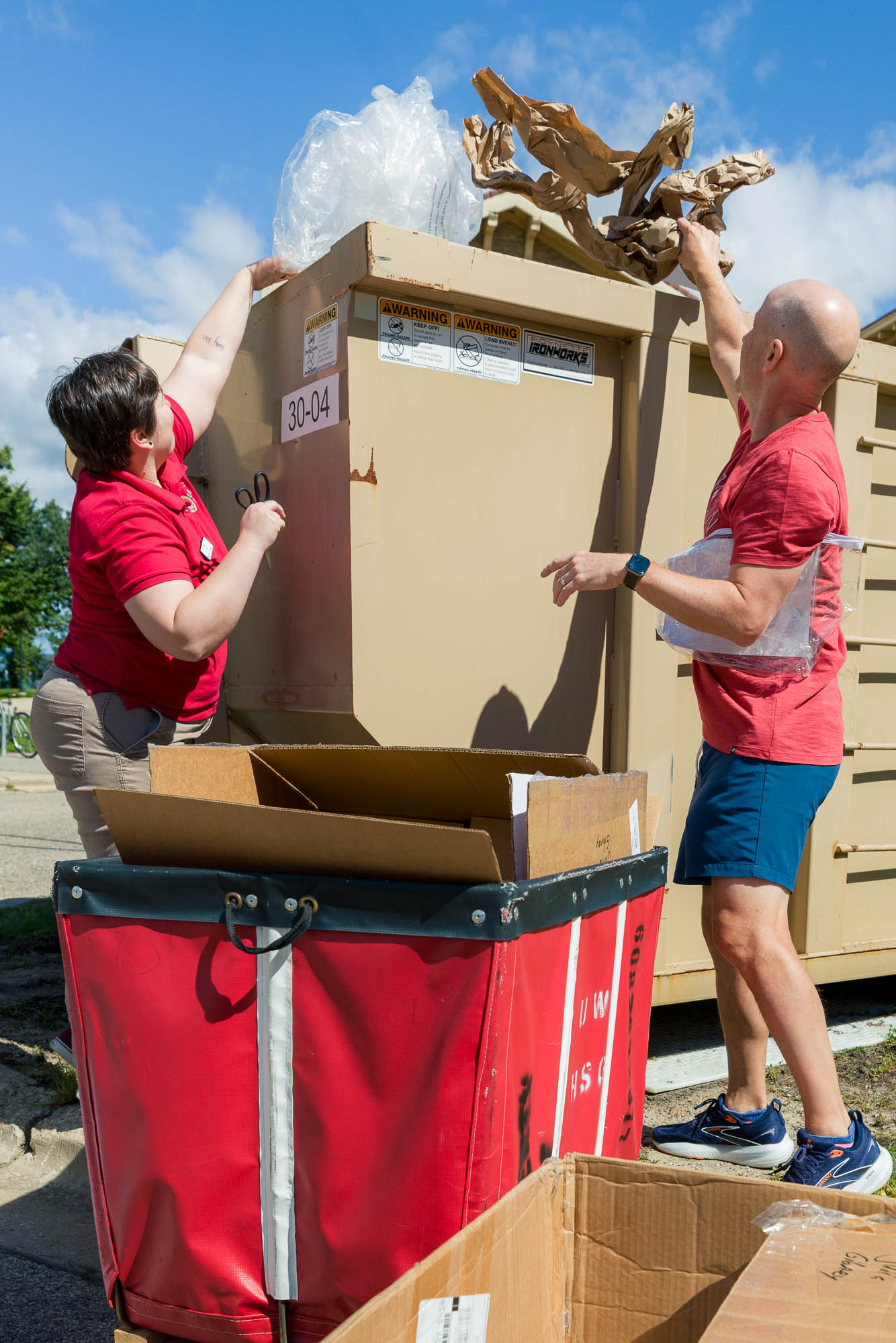 Two people throwing plastic and paper into a dumpster for recycling