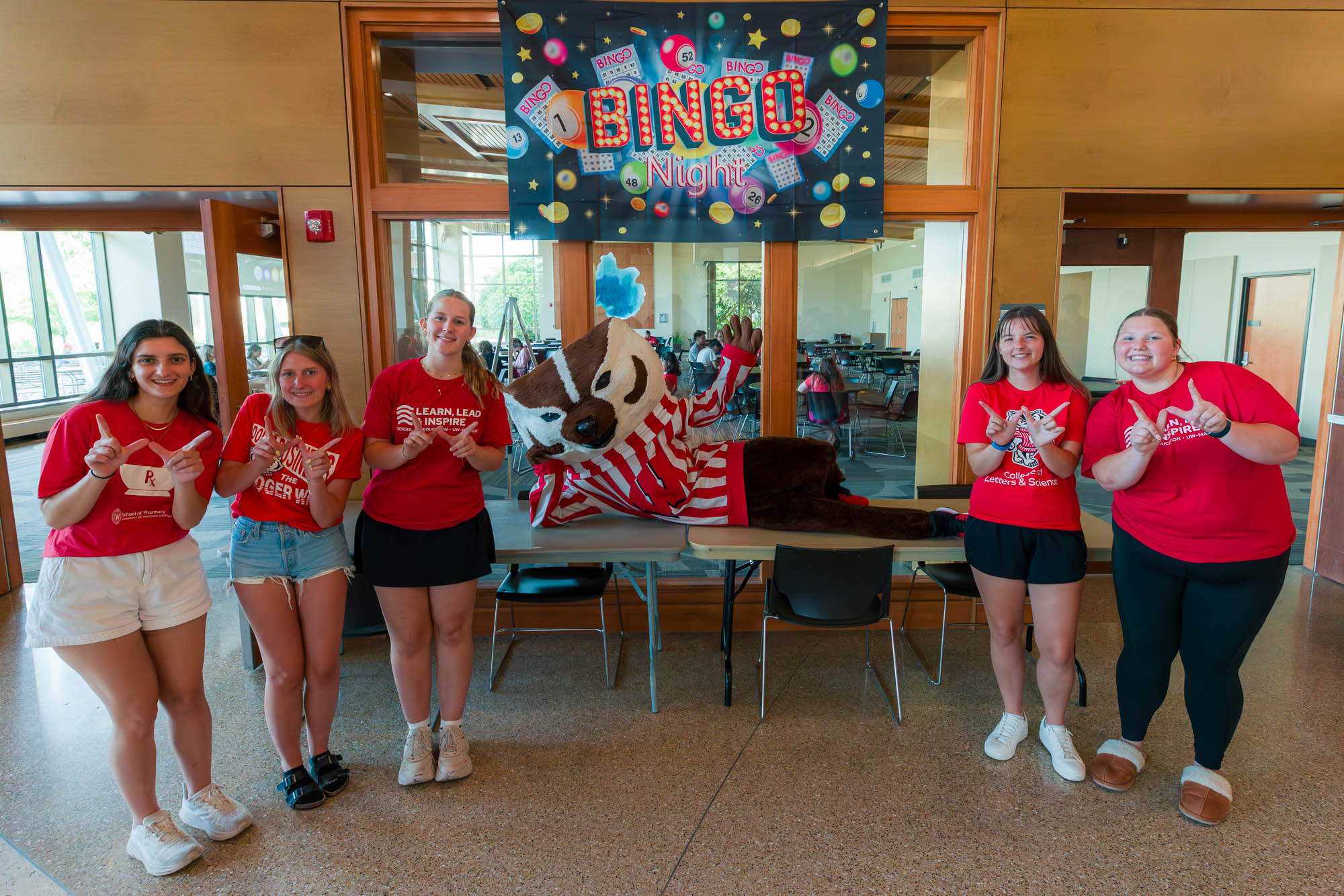 Five people make the letter "W" with their hands while Bucky Badger lays on tables under a sign than reads "Bingo Night"