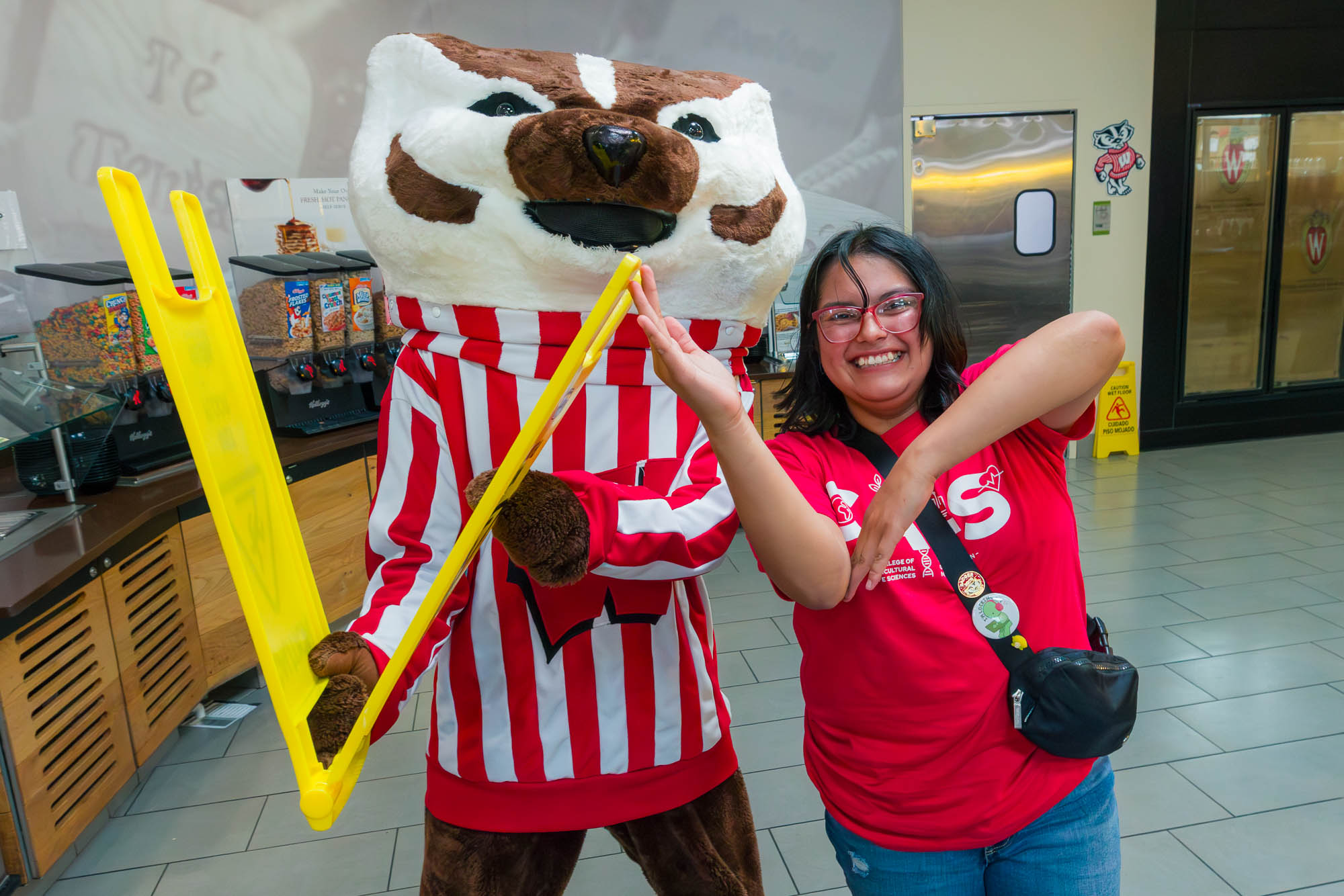 A person and Bucky Badger form a "W" with a yellow "wet floor" sign and their hands