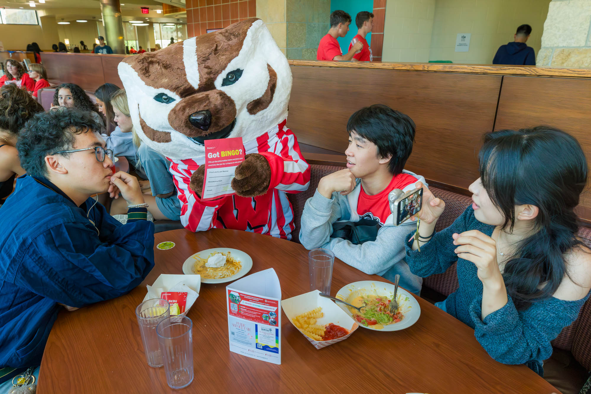 Three people sit with Bucky Badger while eating as Bucky points to a card reading "Got bingo?"