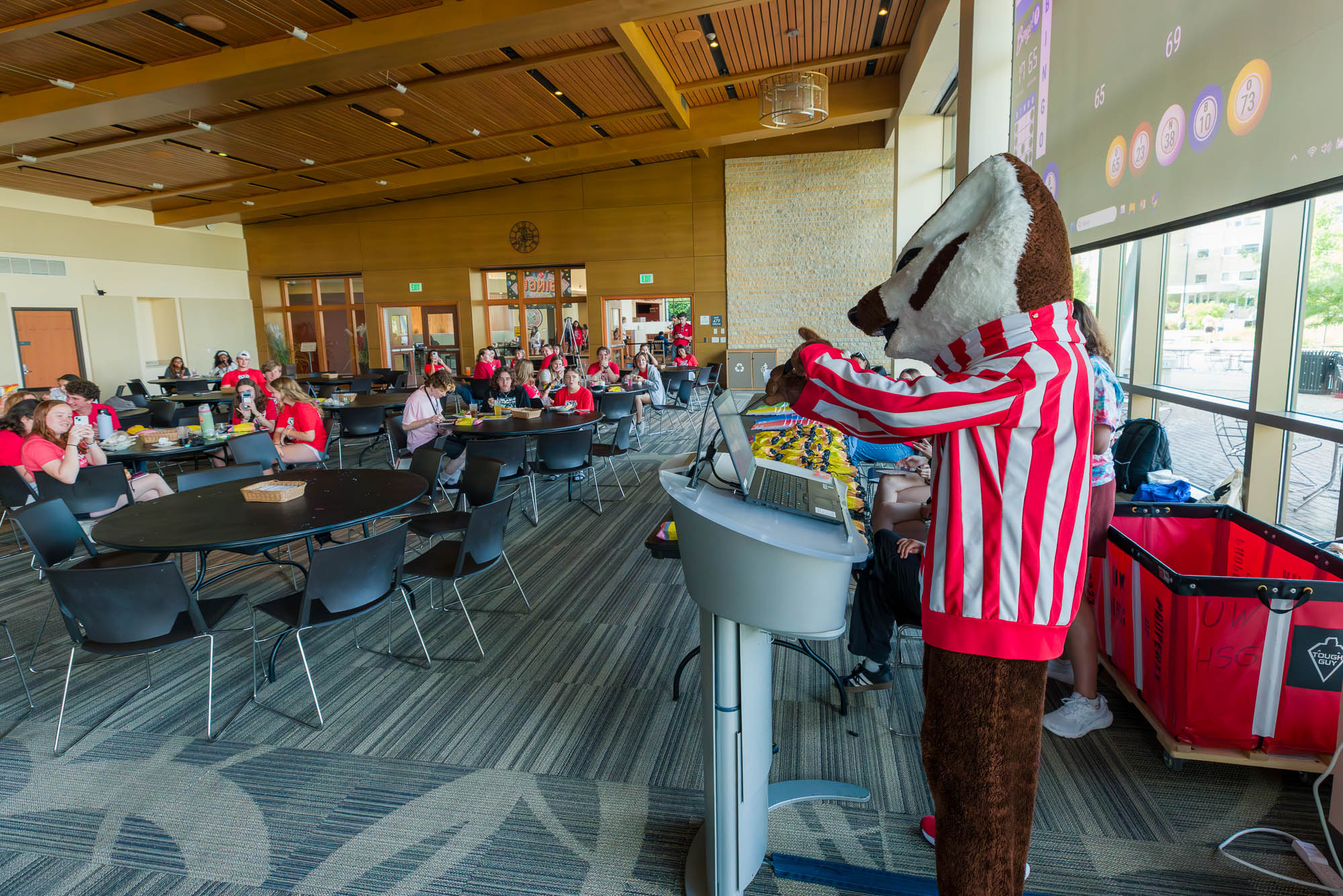 Bucky Badger stands in front of a podium and gestures to other people