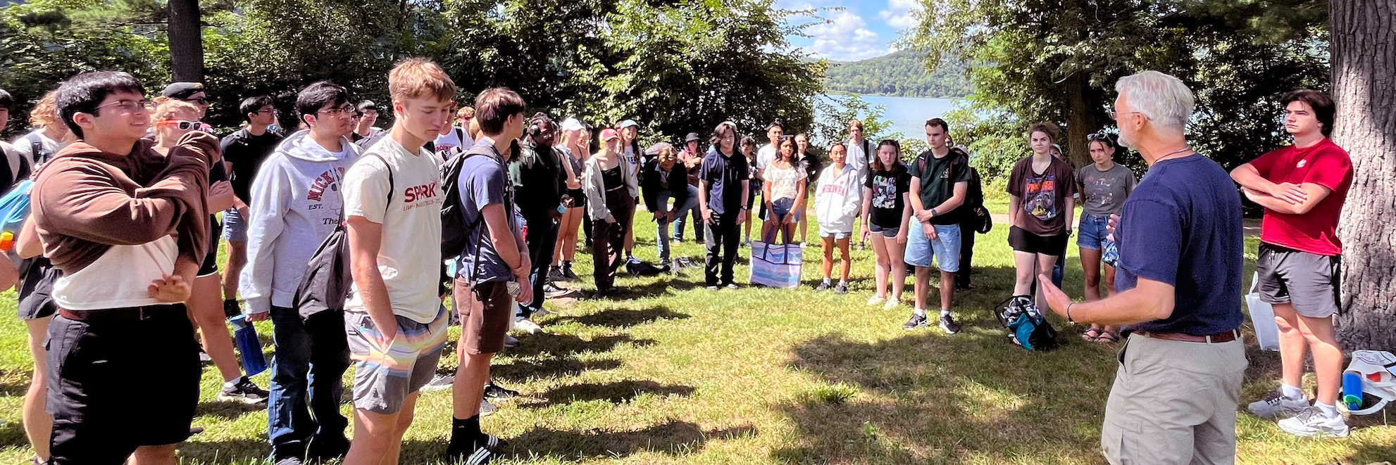 A group of people listen to another person talking with trees and grass around them and a lake in the background