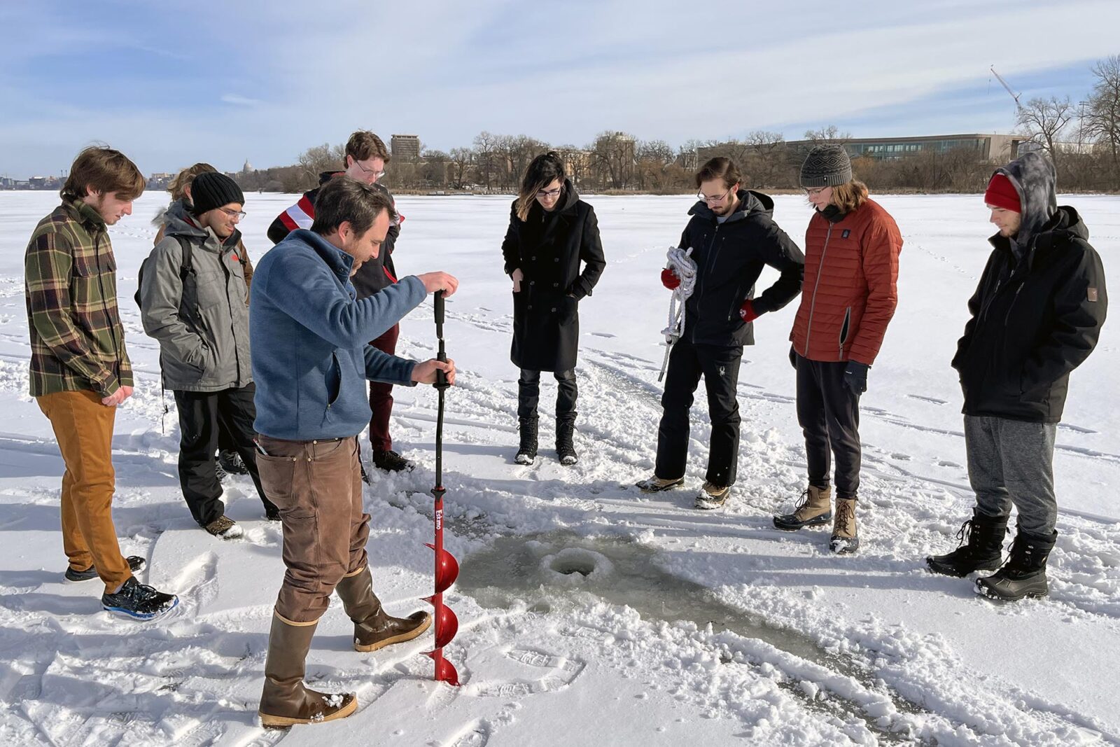A group of people gather around a fresh hole in Lake Mendota for ice fishing, with one person holding an ice auger.