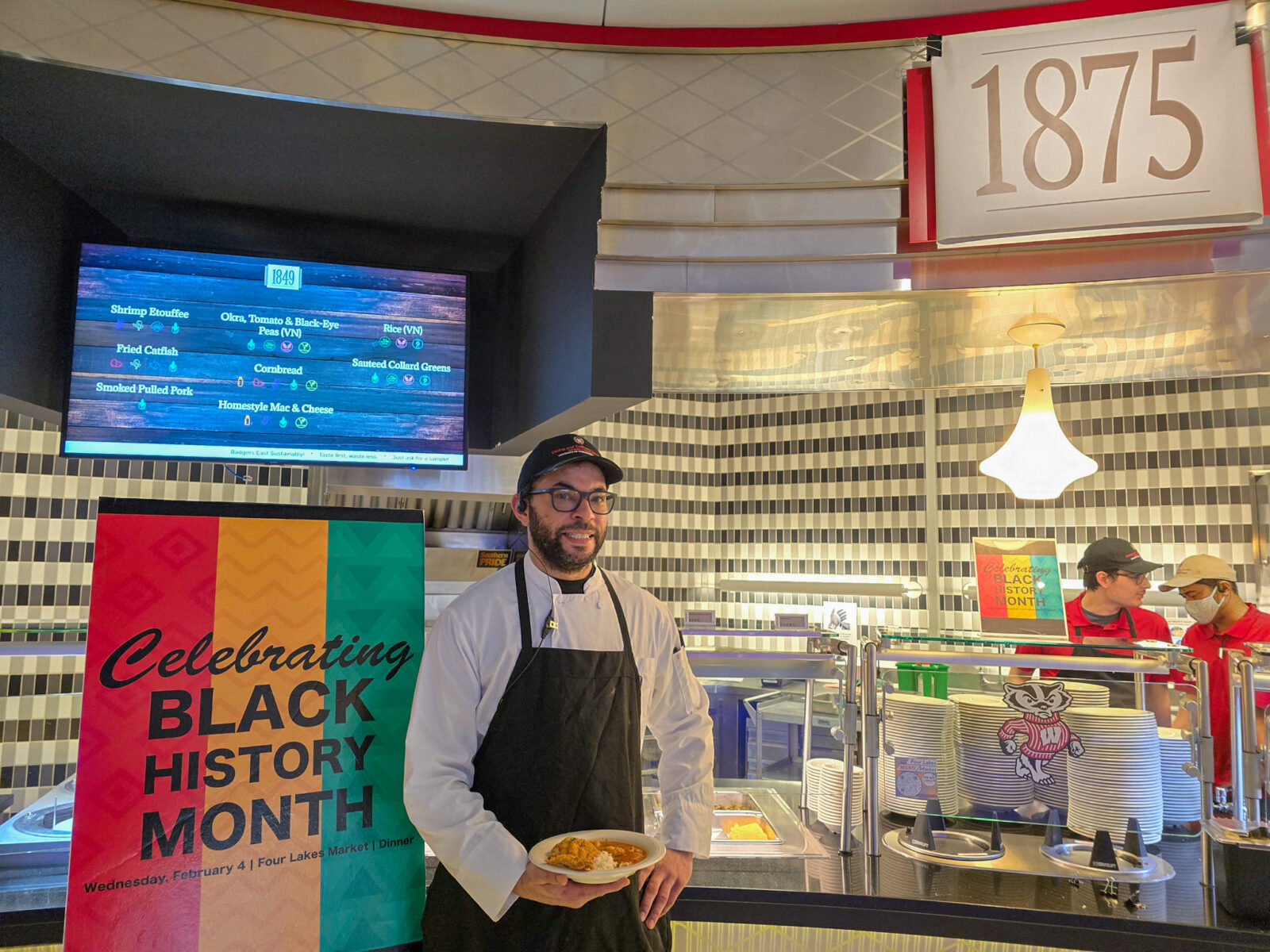 Chef Yusuf posing with a plate and sign at "1875" station for his Black History Month Dinner