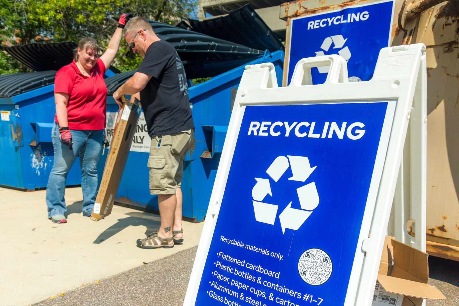 Two people putting cardboard into a container in the background with a sign reading "Recycling" in the foreground