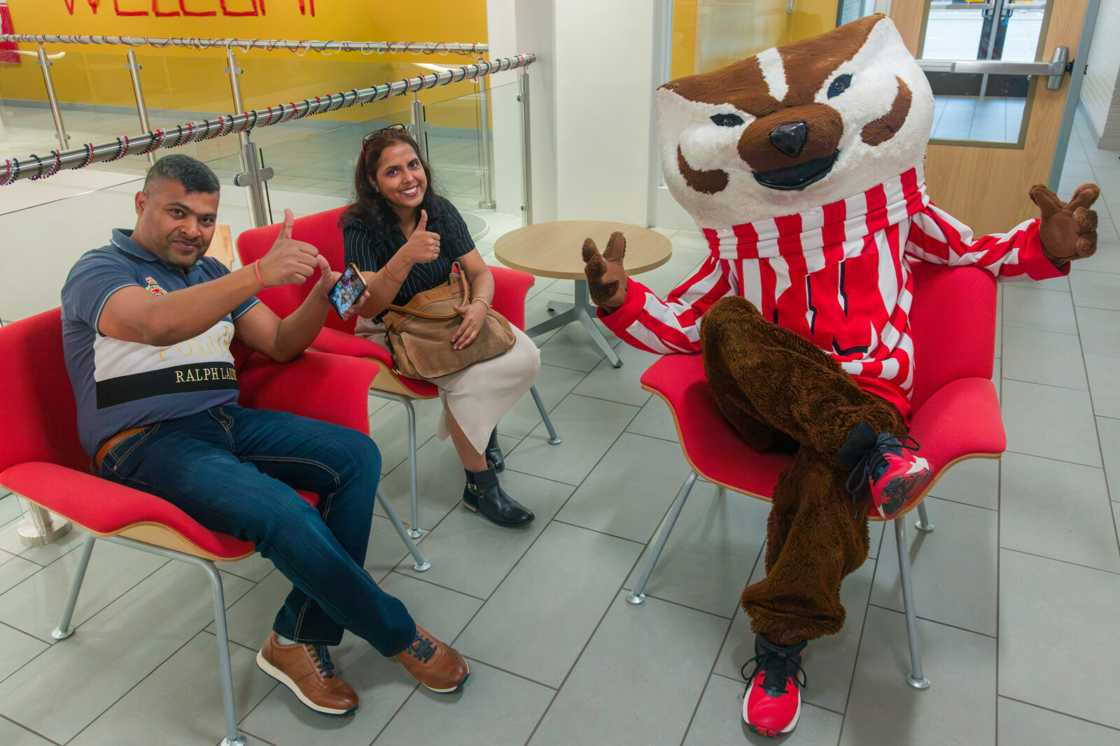 Bucky Badger poses while sitting down with two parents who are giving the thumbs up sign with their hands