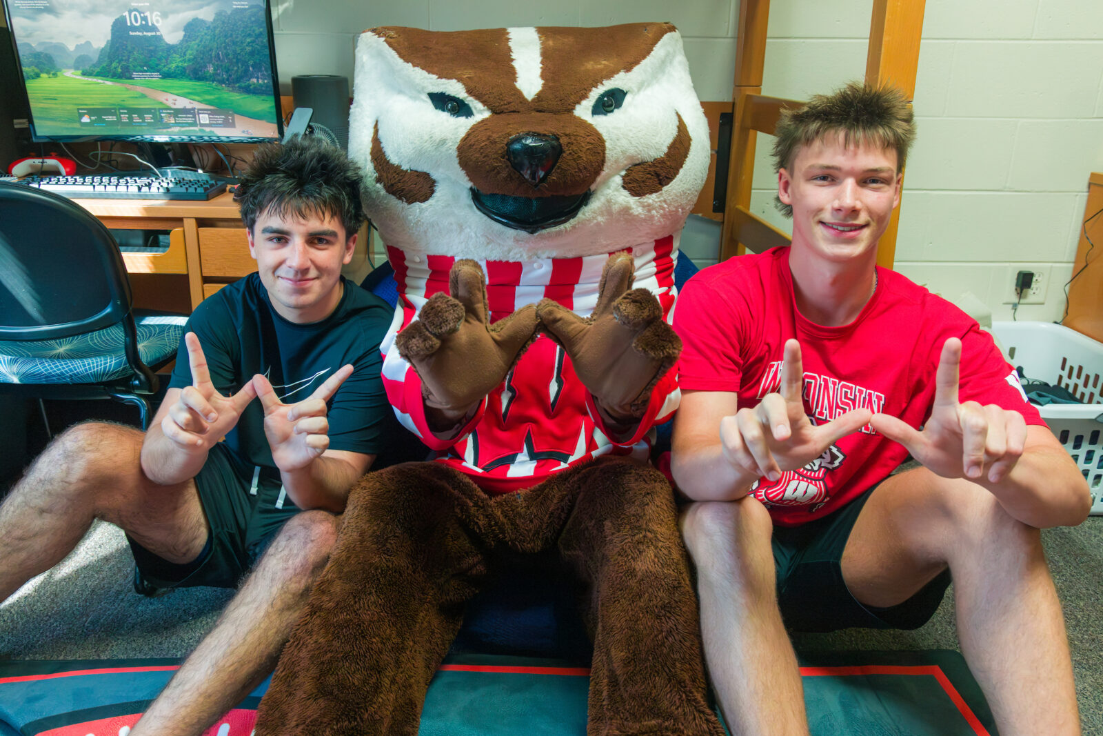 Bucky Badger poses with two people sitting on the floor while making the letter "W" with their hands