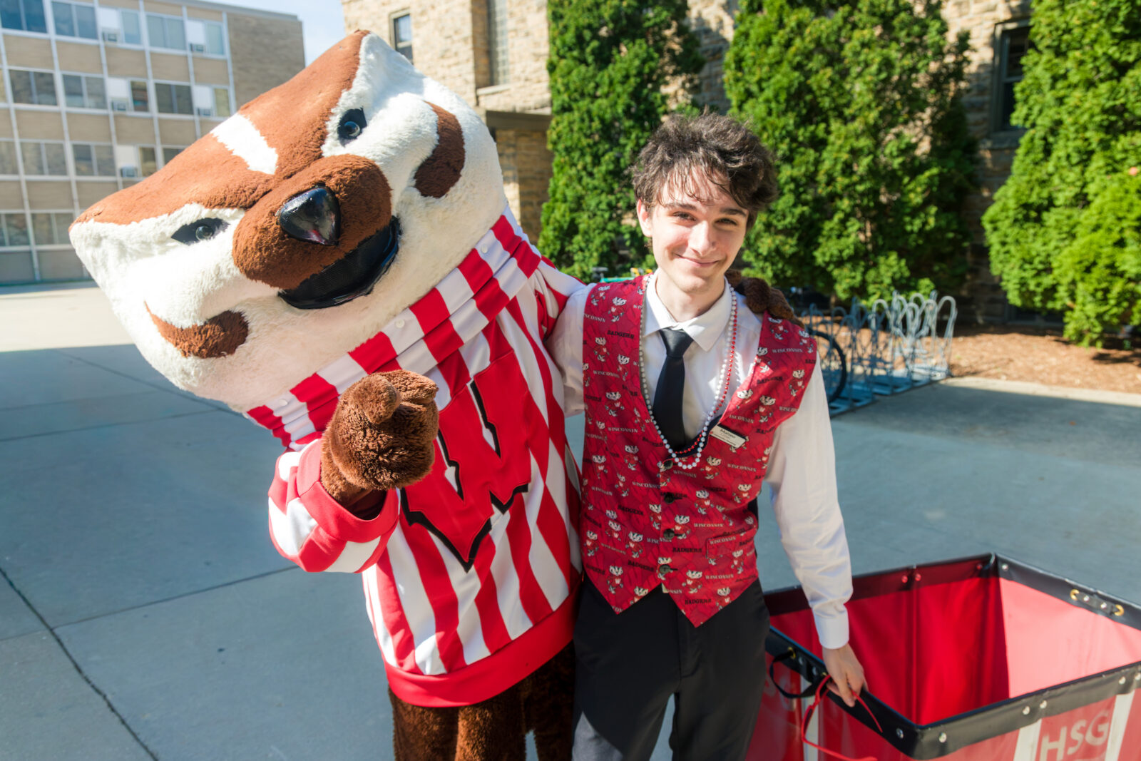 Bucky Badger poses while pointing at the camera with a person wearing a red Bucky vest