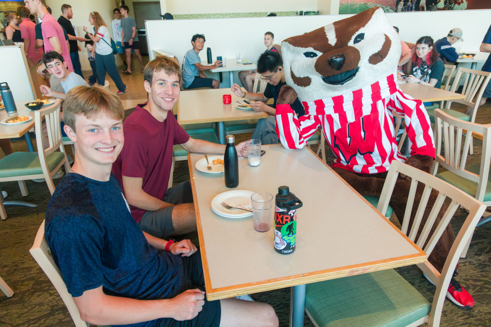 Bucky Badger poses with two people while sitting at a table in a dining room