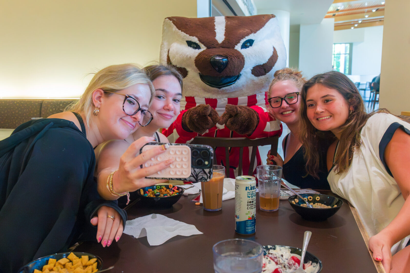 Bucky Badger points at a phone while a group of four people take a selfie with him at a table in a dining room