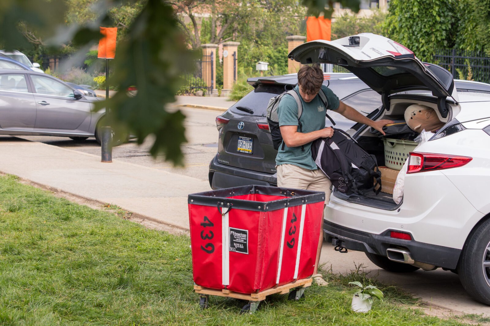 A person unloading a backpack from the tailgate of a vehicle into a red moving cart with green leaves in the foreground