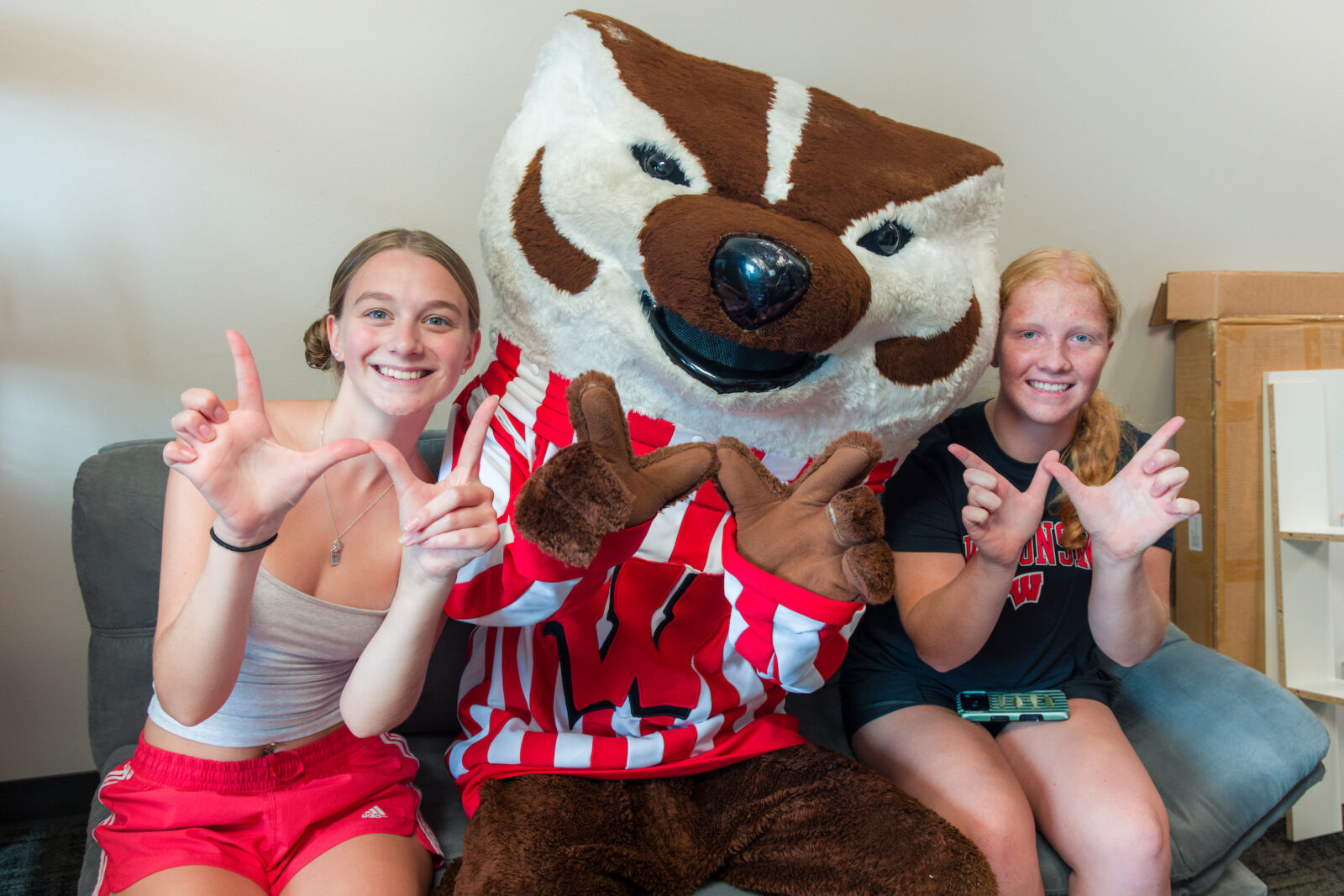 Bucky Badger and two students pose and make the letter "W" with their hands while siting on a futon