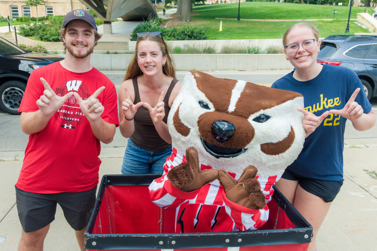 Bucky Badger sits in a red moving cart while posing with three people by making the letter "W" with their hands
