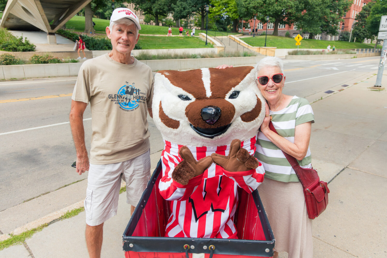 Bucky Badger makes the letter "W" with his hands while sitting in a red moving cart with two people standing next to him