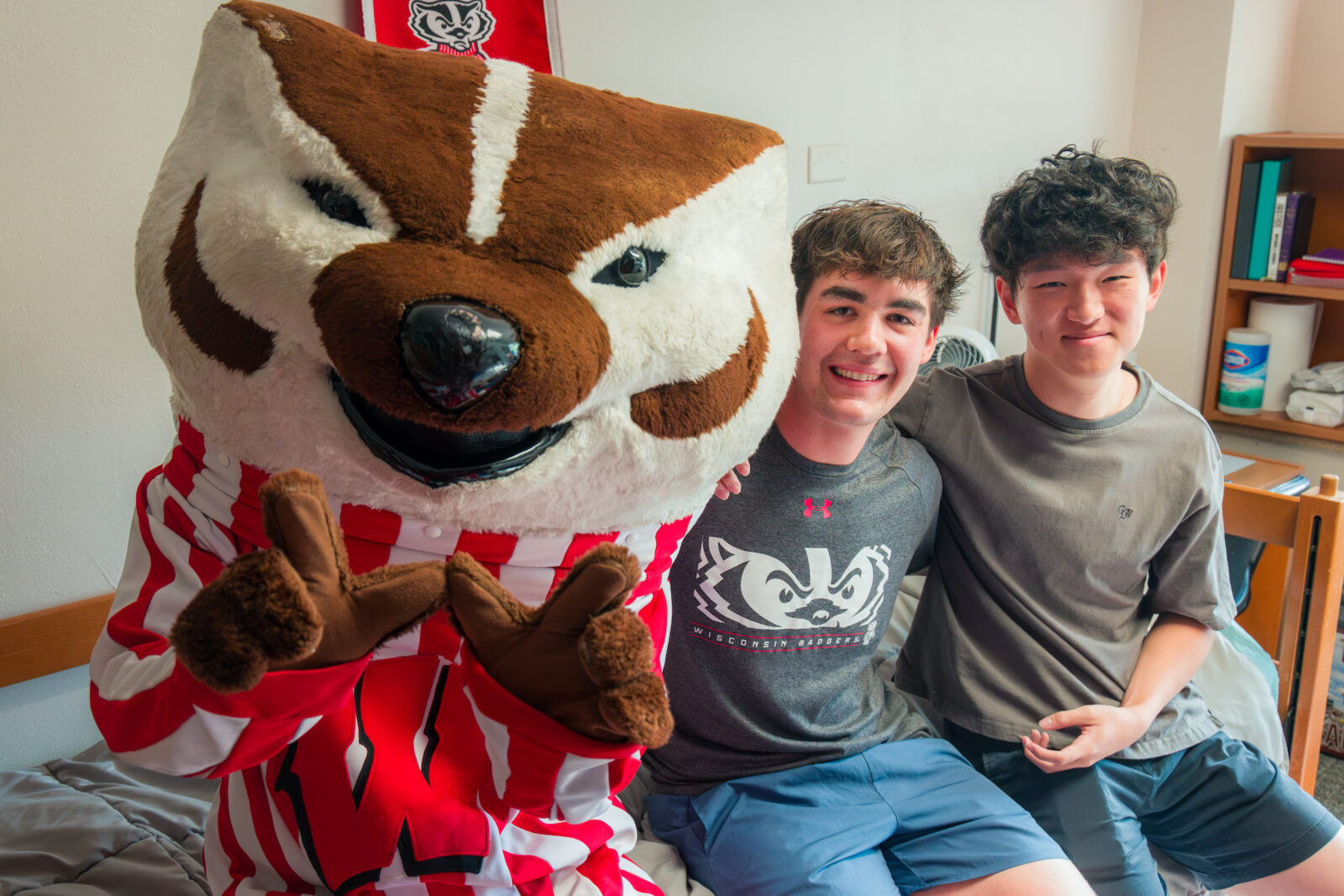 Bucky Badger makes the letter "W" with his hands while sitting on a bed posing with two students