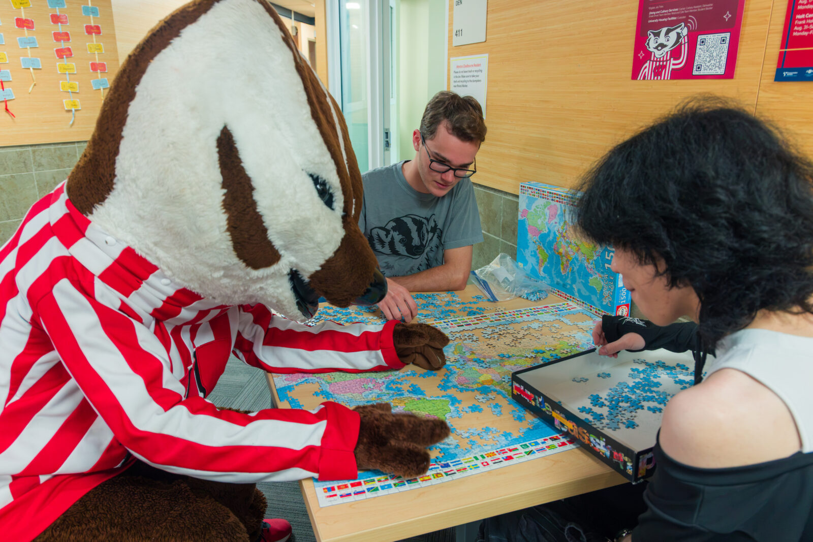 Bucky Badger sits at a table and works on a map of the world puzzle with two students