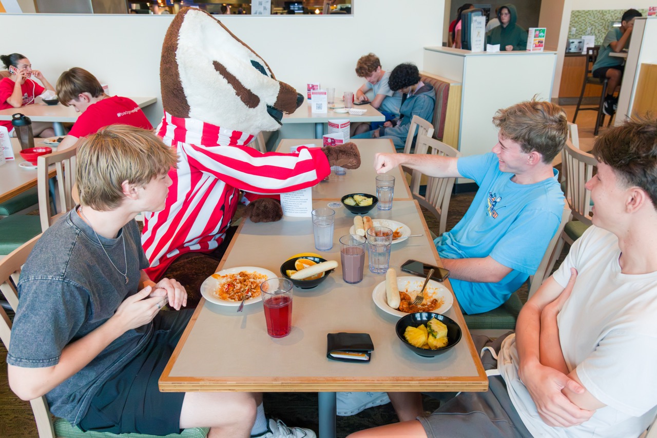 Bucky fist bumping a student at a table in Four Lakes at the Badger Welcome Dinner with two other students