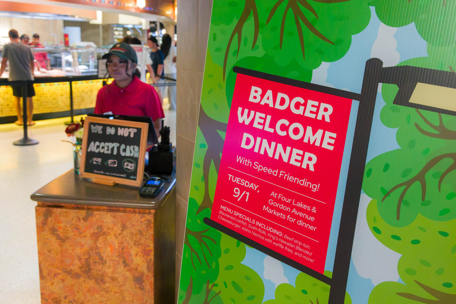 A cashier staff member waits in the background while a sign reading "Badger Welcome Dinner" is pictured in the foreground