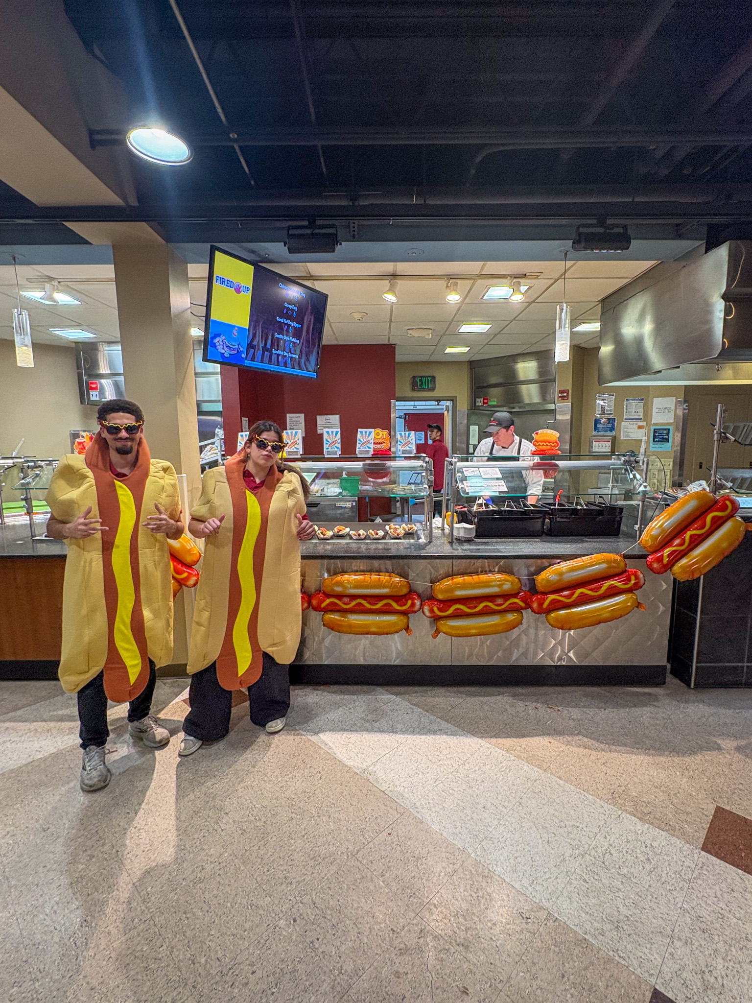 Two student staff dressed up in hot dog costume pose for photo by decorated hot dog station